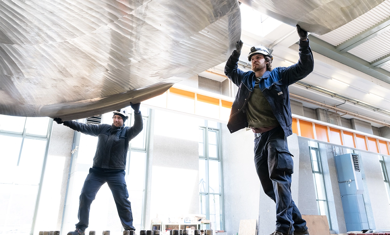 We see two VERBUND employees working on a turbine. Both are wearing work clothes and helmets. The hydroelectric power station can be seen in the background.