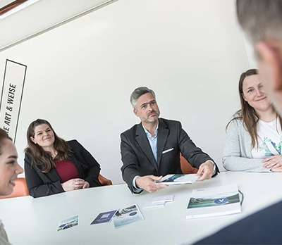 We see three VERBUND recruiters sitting at a table. On the right and left are two more people who are cut off by the camera angle. All the people are wearing business attire and smiling at each other.