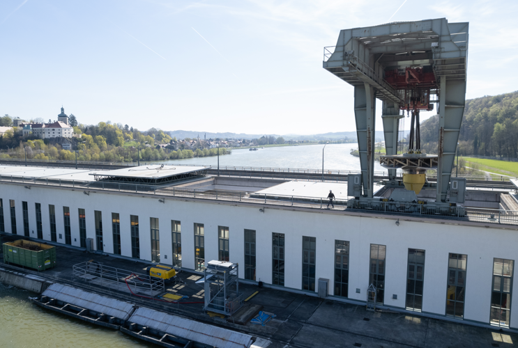 Von einem leicht erhöhten Standpunkt wurde die Fotografie des Krafwerks bei Ybbs gemacht. Wir sehen einen Wasserkraftkran und die Donau bei Sonnenschein. Ein VERBUND-Mitarbeiter ist ganz klein neben dem Kran zu sehen.