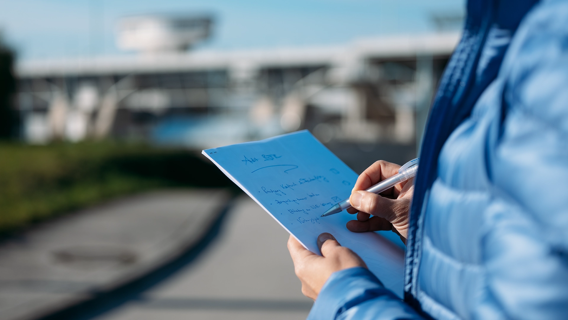 We look directly over the shoulder of a VERBUND employee who is taking notes. He is standing near a hydroelectric power plant, which can be seen very blurred in the background.